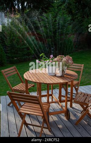 Fleurs en fleurs dans des pots blancs sur une table en bois, terrasse dans le jardin verdoyant, en arrière-plan un arroseur arrose la pelouse et les conifères verts, les jets d'eau brillent dans le contre-jour du soleil du soir Banque D'Images