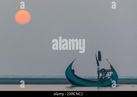 Un bateau de lune traditionnel (Shander Nouka) qui revient sur terre après avoir traversé les vagues à l'extérieur de Cox's Bazar au Bangladesh. Ces bateaux de pêche en forme de lune portent généralement un équipage de 3 à 5 et sont réputés pour leur belle artisanat. Leur forme les rend parfaits pour traverser les vagues pour se rendre au parc de pêche. La forte pollution par les particules de la production de briques tout le long du delta rend le ciel brumeux avec le smog et le soleil orange. Banque D'Images