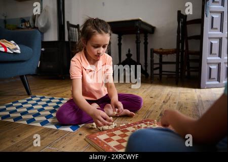 Une jeune fille est absorbée dans une partie d'échecs avec un ami à la maison. L'image capture la mise au point, la concentration et le jeu occasionnel en intérieur. Banque D'Images