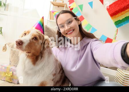 Jeune femme et chien berger australien mignon dans des chapeaux de fête prenant selfie à la maison. Célébration d'anniversaire Banque D'Images