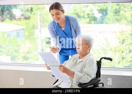 Femme senior en fauteuil roulant avec infirmière lisant le journal près de la fenêtre à la maison Banque D'Images