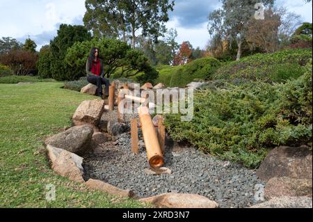 Vue en angle bas d'une pièce d'eau en bambou menant à une touriste, en réflexion, assise sur un rocher près d'une petite cascade dans un jardin japonais. Banque D'Images