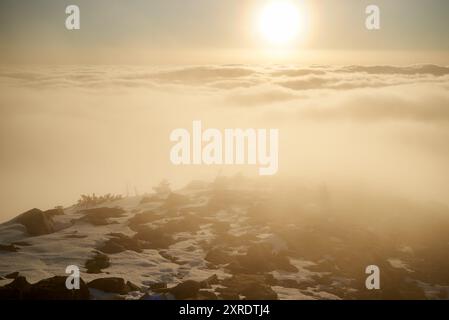 Lever du soleil sur le sommet de montagne enneigé. La lumière dorée chaude du soleil levant baigne les nuages dans des tons doux d'orange et de rose. La neige et les roches dispersées au premier plan ajoutent de la texture et de la profondeur à la scène. Banque D'Images