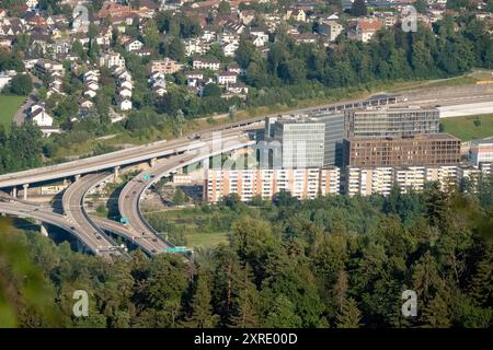 Autobahn A3 und Stadtteil Wollishofen Das Bild zeigt einen detaillierten Blick vom Uetliberg auf die Autobahn A3, die sich durch den Zürcher Stadtteil Wollishofen schlängelt. Die Autobahn ist eine wichtige Verkehrsader, die Zürich mit anderen Teilen der Schweiz verbindet. IM Vordergrund ist ein markantes Autobahnkreuz zu sehen. Dahinter erstreckt sich ein modernes Bürogebäudeensemble, das durch Seine klare, rechteckige Architektur auffällt. DAS Gebiet ist von dicht bewaldeten Hügeln umgeben, die dem städtischen Umfeld eine Grüne und natürliche kulisse verleihen. IM Hintergrund sind Wohngebiete Banque D'Images