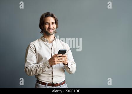 Jeune homme d'affaires heureux tenant le téléphone portable debout isolé sur le mur gris. Banque D'Images