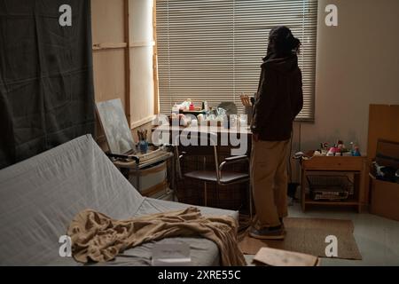 Femme debout dans une chambre confortable regardant par la fenêtre avec une expression réfléchie, chambre remplie d'objets personnels sur le bureau et le lit soigneusement fait avec une couverture posée sur le dessus Banque D'Images