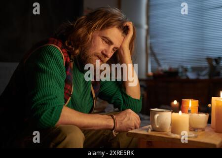 Homme barbu avec de longs cheveux assis à une table éclairée aux chandelles avec plusieurs tasses, portant un pull vert et une chemise rayée, apparaissant introspective. Ombres projetées à la flamme dans un cadre confortable Banque D'Images