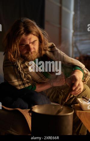 Homme barbu avec de longs cheveux remuant le pot sur la chaleur dans un cadre intérieur rustique avec la lumière du jour se versant à travers la fenêtre, s'engageant franchement dans le processus de cuisson Banque D'Images