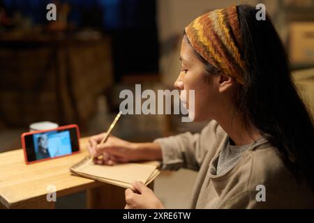 Jeune femme dessinant tout en regardant un tutoriel d'art en ligne à l'aide d'un crayon en bois et d'un carnet de croquis, axé sur l'apprentissage et le dessin dans un cadre intérieur confortable Banque D'Images