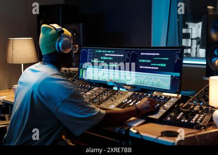 Producteur de musique afro-américain travaillant avec des équipements de mixage dans un studio bien éclairé rempli de divers appareils audio, portant des écouteurs et axé sur les moniteurs Banque D'Images