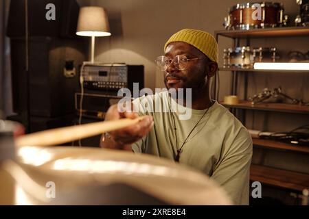 Homme jouant de la batterie dans un studio d'enregistrement professionnel, montrant la concentration et la passion portant un bonnet jaune, des lunettes. L'arrière-plan comprend l'équipement audio, les ensembles de batterie, les étagères Banque D'Images