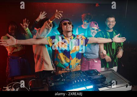 Groupe de jeunes gens excités dans des vêtements à la mode gardant leurs bras levés tout en dansant derrière un heureux mâle deejay debout près de la table d'harmonie Banque D'Images