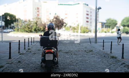 Un homme sur une moto roule dans une rue avec un bâtiment en arrière-plan. L'homme porte un casque et une chemise noire Banque D'Images