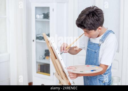 Un jeune garçon peint un tableau avec un pinceau et une palette. Il porte une chemise blanche et une salopette bleue Banque D'Images