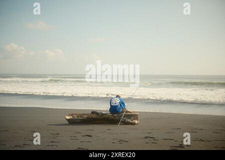 Tabanan, Bali, Indonésie - 9 août 2024 : un homme pêche ou fait du surf assis sur une bûche sèche au bord d'une plage de sable noir Banque D'Images