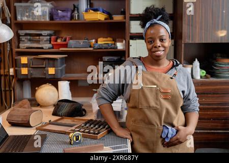 Jeune femme debout devant l'établi avec divers outils et fournitures, tenant un morceau de tissu et souriant avec confiance dans un atelier de bois rempli de matériaux d'artisanat Banque D'Images