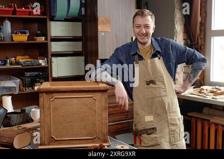 Artisan masculin souriant portant tablier travaillant sur armoire en bois en atelier entouré d'outils et de matériaux créant le projet de menuiserie Banque D'Images