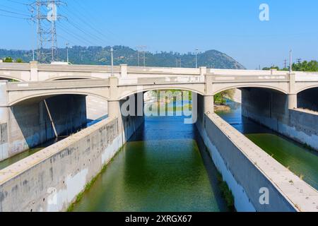 Vue sur la rivière Los Angeles qui coule sous un pont, avec des canaux en béton et de l'eau verte, entourée de collines et de lignes électriques Banque D'Images