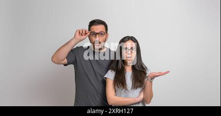 Portrait d'un jeune couple inquiet et effrayé habillé en décontractés regardant la caméra de façon choquante. Femme étonnée faisant des gestes et homme tenant des lunettes pendant s Banque D'Images