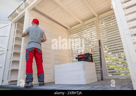 Un homme en pantalon rouge organise des étagères dans un hangar en bois, se concentrant sur l'organisation tout en profitant de la lumière du soleil en plein air. Banque D'Images