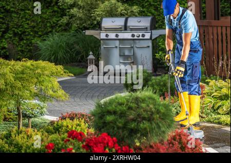 Un jardinier en bottes jaunes coupe les buissons dans une cour magnifiquement aménagée avec des fleurs colorées et un grill. Banque D'Images