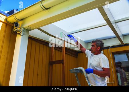 Vue rapprochée de l'homme peignant le cadre de patio blanc avec rouleau à sa maison. Banque D'Images