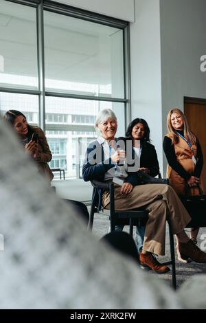Femme d'affaires mature parlant lors d'une conférence, s'engageant avec un public souriant et diversifié dans un cadre de bureau moderne. Banque D'Images