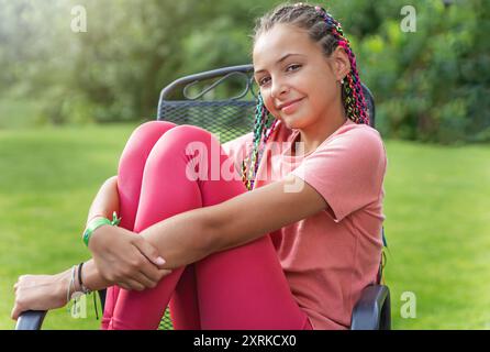 Vue de côté de la jeune fille avec des tresses colorées dans ses cheveux pose dans des vêtements d'action assis sur la chaise de jardin en métal à l'extérieur. Horizontalement. Banque D'Images