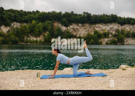 Une femme pratiquant élégamment une pose de yoga au bord d'un lac serein, entourée par la beauté de la nature Banque D'Images