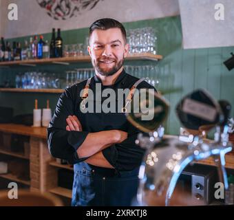 Portrait d'heureux barman barbu souriant vêtu d'un uniforme noir avec un tablier au comptoir de bar avec des robinets de bière pression. Des gens qui réussissent, un travail acharné, Banque D'Images