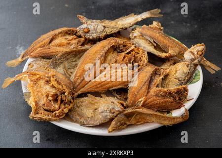 Ikan masin goreng ou poisson salé frit dans une plaque blanche, isolé sur fond noir. Cuisine traditionnelle malaisienne et indonésienne. Banque D'Images