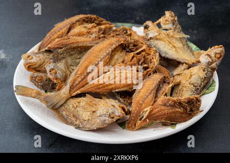 Ikan masin goreng ou poisson salé frit dans une plaque blanche, isolé sur fond noir. Cuisine traditionnelle malaisienne et indonésienne. Banque D'Images