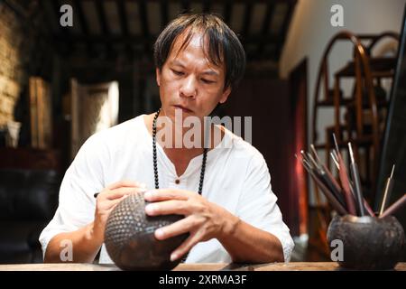 (240811) -- HAIKOU, 11 août 2024 (Xinhua) -- Wu Mingju sculpte une coquille de noix de coco dans un atelier à Haikou, dans la province de Hainan, dans le sud de la Chine, le 9 août 2024. La sculpture de coquille de noix de coco, une forme d'art qui implique la création de dessins à l'aide d'outils de sculpture sur la coquille dure d'une noix de coco, a été répertoriée comme l'un des patrimoines culturels immatériels nationaux en 2008. Wu Mingju, un héritier de la sculpture de coquille de noix de coco, s'est engagé à l'héritage et à la promotion de cet art ancien et complexe depuis 33 ans en explorant l'innovation technique, en mettant en place des cours de sculpture de coquille de noix de coco dans les écoles et la cultivatine Banque D'Images