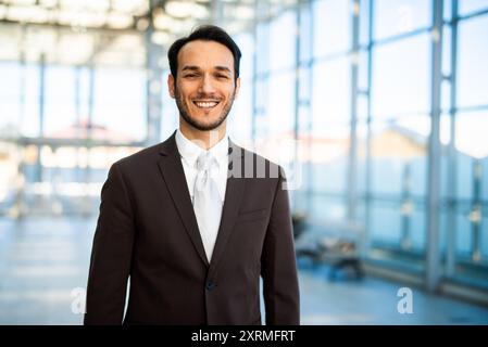 Jeune homme d'affaires portant un costume se tient debout dans un immeuble de bureaux moderne avec de grandes fenêtres, à l'air heureux et réussi Banque D'Images