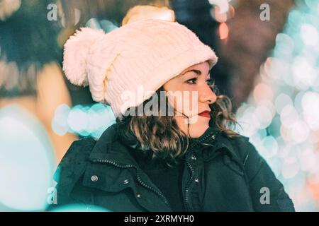 Femme blonde aux cheveux bouclés avec chapeau blanc regardant sur le côté avec des lumières de Noël sur fond flou Banque D'Images