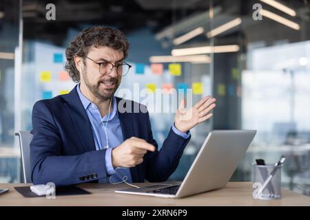 Homme d'affaires confiant dans des lunettes conduisant la réunion virtuelle sur ordinateur portable avec téléphone. Engagé dans la communication en ligne, les mains gestuelles expressives. Environnement de bureau moderne Banque D'Images