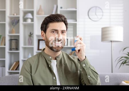 Homme utilisant un inhalateur pour soulager l'asthme assis à la maison souriant. Personne démontrant l'utilisation appropriée d'un dispositif médical pour des troubles respiratoires. Concept de soins de santé, thérapie respiratoire, maladie chronique. Banque D'Images