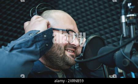 Homme souriant avec des lunettes portant des écouteurs dans un studio d'enregistrement insonorisé, prêt pour podcast ou session vocale. Configuration professionnelle avec microphone et filtre pop, fond de mousse acoustique. Banque D'Images