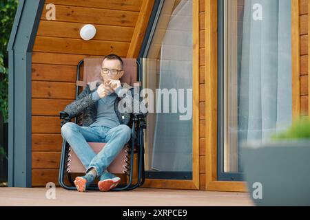 Homme d'âge moyen allume un cigare tout en étant assis dans une chaise longue sur la terrasse d'une maison de campagne Banque D'Images