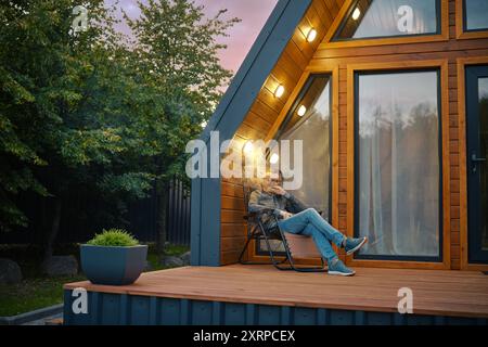 L'homme d'âge moyen aime un cigare tout en étant assis sur la terrasse de la cabane en rondins moderne dans la soirée Banque D'Images