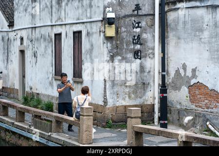 Suzhou, Chine - 11 juin 2024 : Un homme prend une photo d'une femme assise sur une balustrade de pierre près d'un canal devant un bâtiment blanc altéré avec Chine Banque D'Images