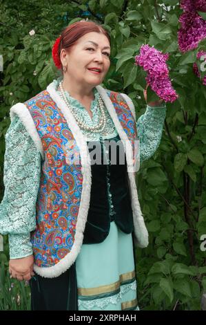 Stanitsa Borgustanskaya, Pyatigorsk, 9 mai 2024. Une femme cosaque âgée en vêtements nationaux près de fleurs de lilas. Cour cosaque. Colonie cosaque Banque D'Images