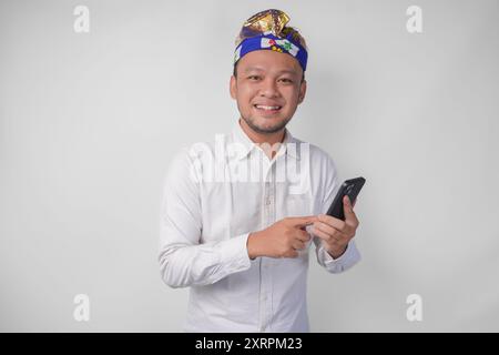 Homme balinais excité portant une chemise blanche et coiffe traditionnelle souriant joyeusement tout en utilisant un smartphone, en répondant un message, en lisant des nouvelles Banque D'Images