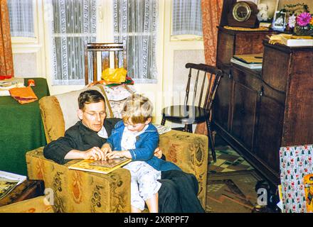 Père et jeune enfant assis dans un fauteuil dans le salon à la maison avec des livres à Noël, Royaume-Uni 1956 Banque D'Images