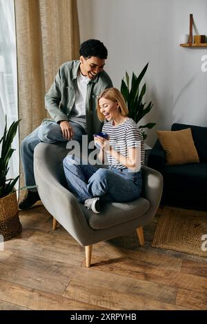 Jeune couple appréciant les uns les autres compagnie dans un appartement moderne. La femme est assise dans un fauteuil gris tandis que l'homme s'appuie contre le dos de celui-ci Banque D'Images