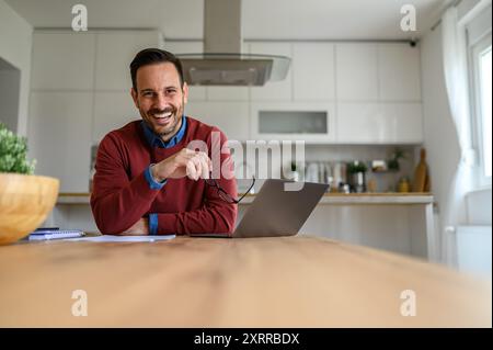Bel homme d'affaires tenant des lunettes et souriant à la caméra avec ordinateur portable sur le bureau dans le bureau à domicile Banque D'Images