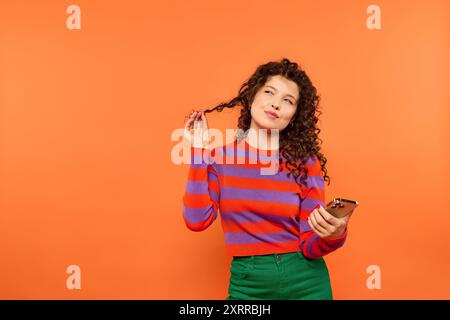 Une jeune femme aux cheveux bouclés pose sur un fond orange vif, tenant son téléphone et regardant soigneusement sur le côté. Banque D'Images