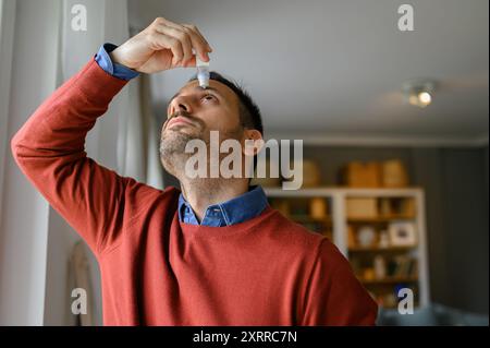 Jeune homme appliquant un lubrifiant pour les yeux pour traiter les yeux secs ou les allergies tout en étant debout à la maison Banque D'Images