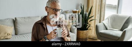 Un homme mûr aux cheveux gris et aux tatouages apprécie une tasse de café tout en travaillant à domicile. Banque D'Images