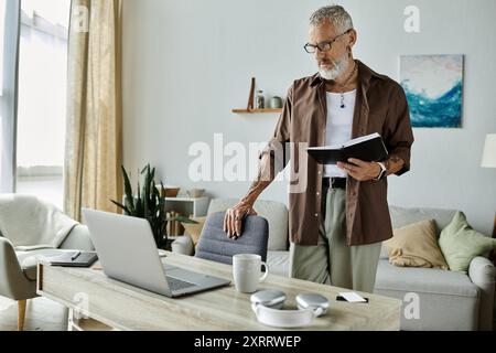 Un homme plus âgé tatoué avec les cheveux gris travaille à la maison, debout près de son bureau avec un cahier, concentré sur son travail. Banque D'Images